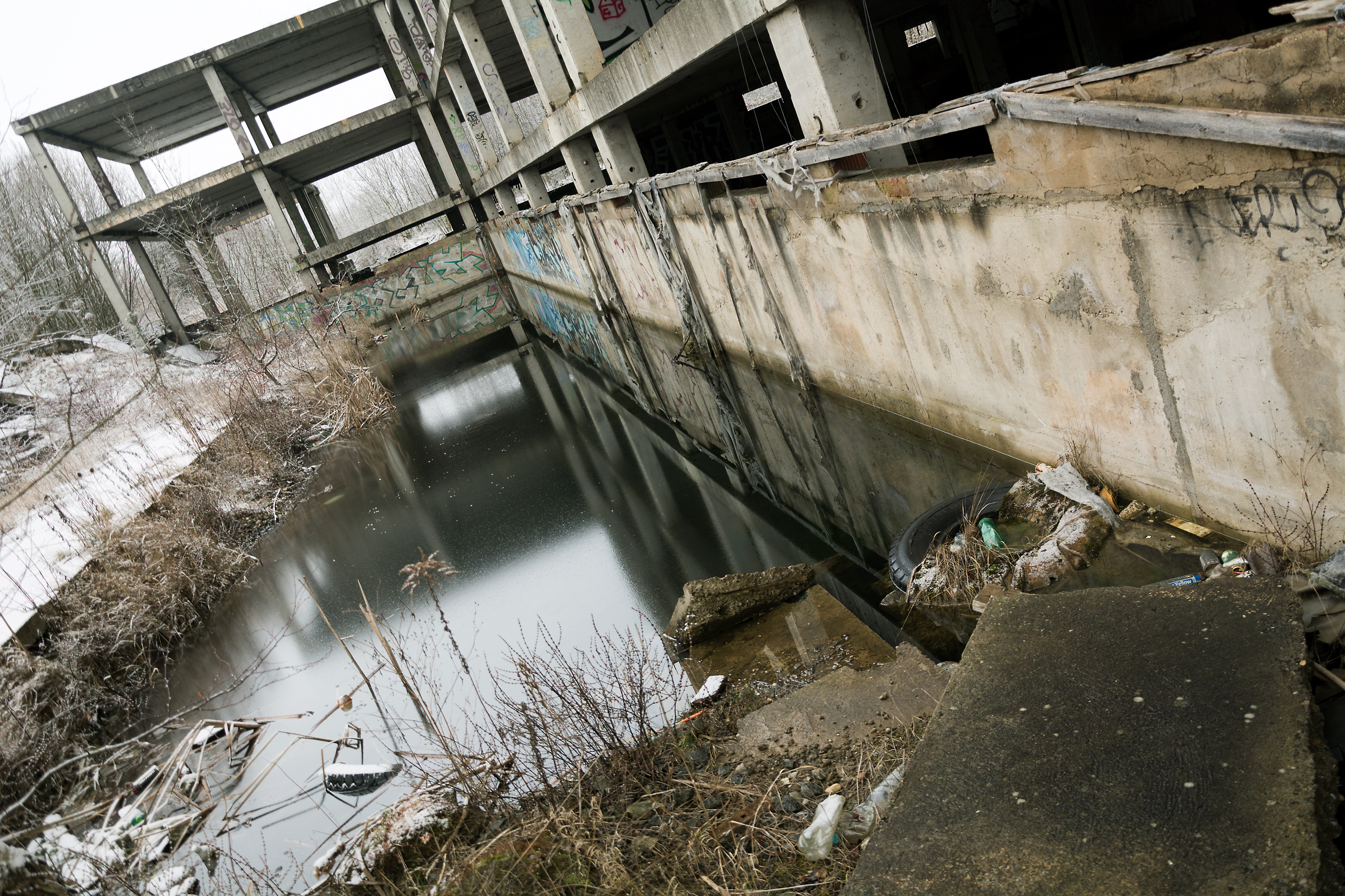 A large concrete building, covered in trash and graffiti, with a large pool of water by it